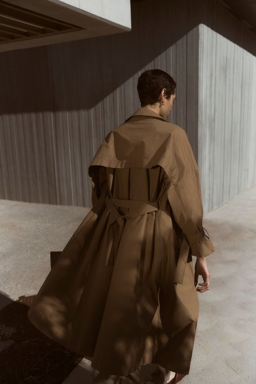 Woman in tan trench coat stands against a corrugated concrete wall, showcasing fashion and architectural elements in muted sunlight.