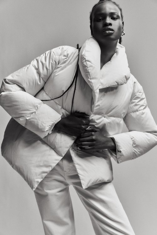 A black woman models a large white quilted puffer jacket and white jeans in a studio with soft lighting, captured in a medium shot.