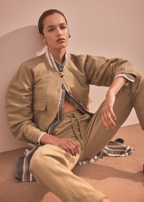 A woman poses in a tan jacket and pants, seated against a wall. The light illuminates her face, highlighting her neutral expression.
