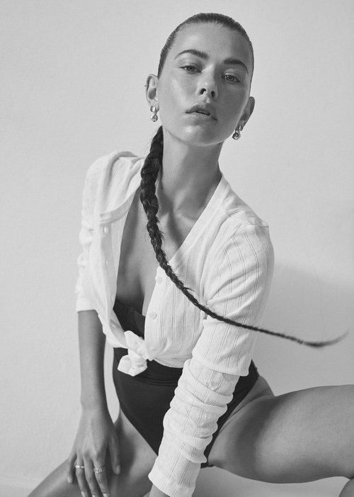 Black-and-white portrait of a young woman with a braided ponytail, posing in a white cardigan and dark bodysuit against a plain backdrop.