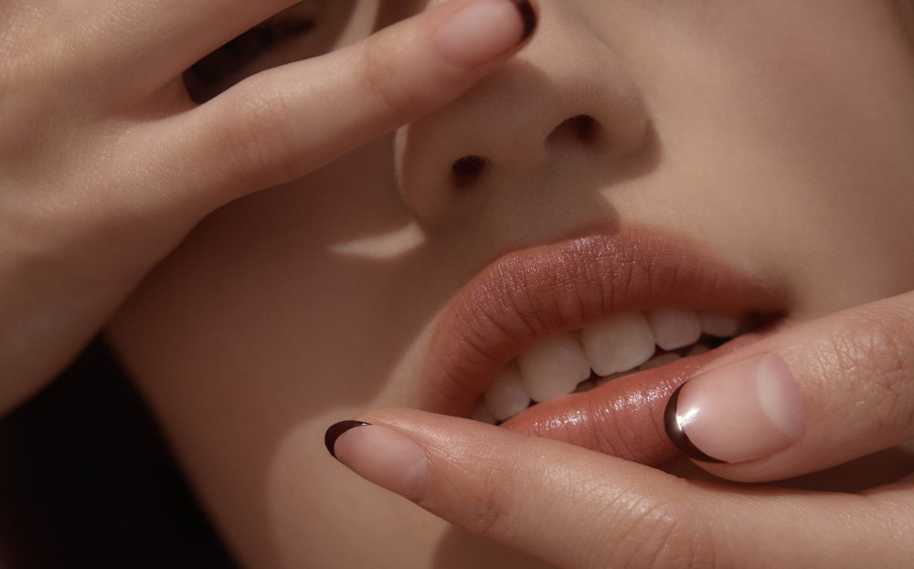 Close-up studio shot of a fair-skinned person with brown lipstick and dark-tipped nails with hands near their face.
