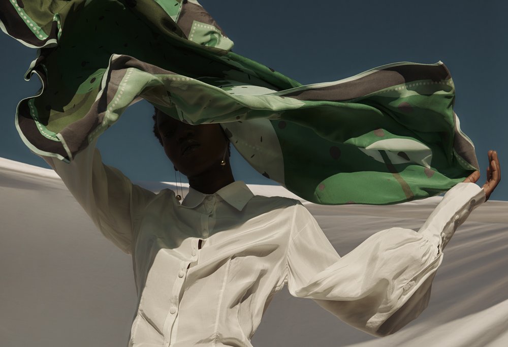 Black model in white shirt holding green patterned fabric against a blue sky, creating an airy fashion portrait with natural light.