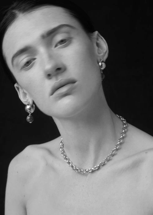 Close-up portrait of a woman wearing silver chain necklace and dangling earrings against a dark background, photographed by Anne Peeters.