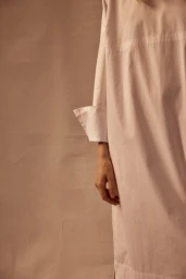 Close-up of a person wearing a white cotton shirt with rolled-up cuffs against a textured neutral background, photographed by Anne Peeters.