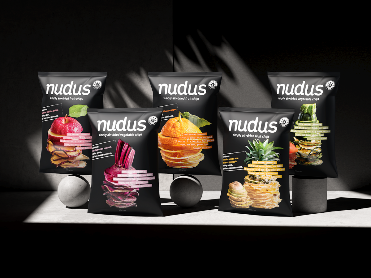 Eye-level studio shot of multiple dark packages of Nudus brand dried vegetable and fruit chips with fresh ingredients displayed.