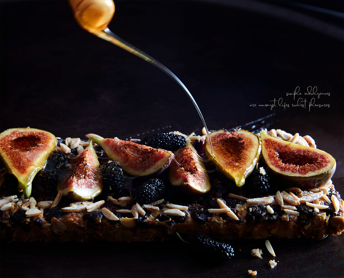 Overhead shot of honey being drizzled on figs and mulberries atop a dessert bar, dark backdrop, food photography, dessert presentation.