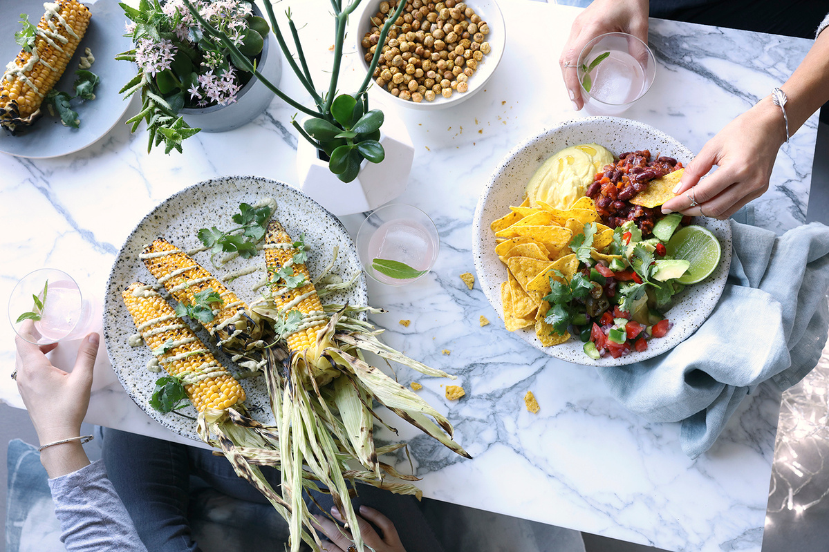 Overhead shot of grilled corn on the cob and a bowl of chips with salsa and toppings on a marble table, alongside drinks and plants.