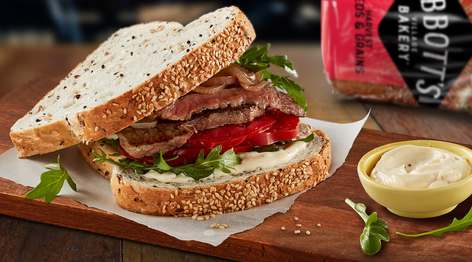Close-up of an open-faced steak sandwich with tomatoes, caramelized onions, greens, and mayo on multigrain bread, served with dipping sauce.