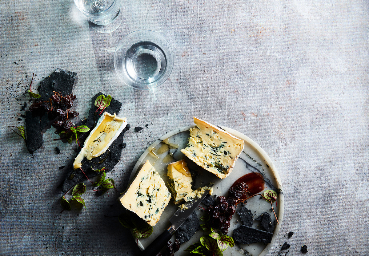 Overhead shot of wedge cheeses on a marble board with black stones, small green leaves, and two glasses of water.