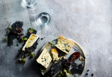 Overhead food photography of blue cheese wedges, dried fruit, and a cheese knife on a round marble board atop a grey concrete surface.