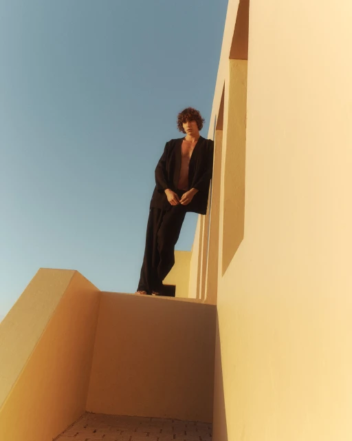 Low-angle shot of a shirtless adult male in a black suit standing on a rooftop against a bright blue sky and light yellow walls.
