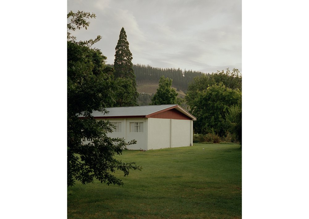 Exterior shot of a white building with a red roof surrounded by green trees and grass under an overcast sky in a rural area.