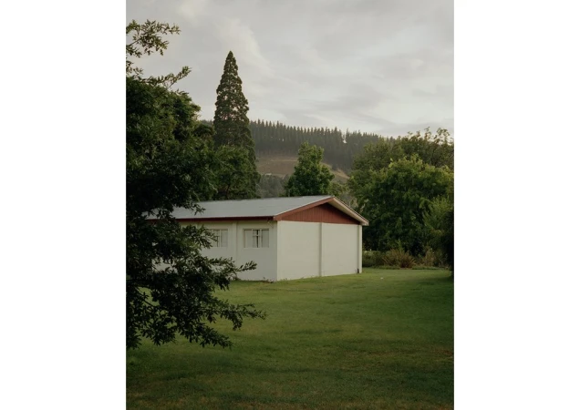 Exterior shot of a white building with a red roof surrounded by green trees and grass under an overcast sky in a rural area.