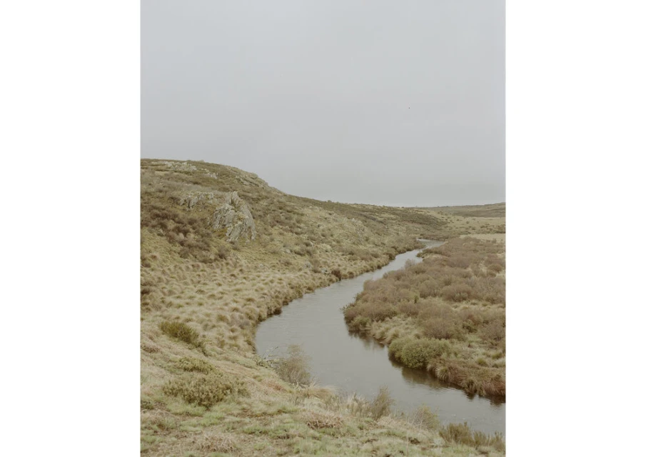 A river winds through a valley landscape under an overcast sky. Green and brown grasses cover the hills. Soft, diffused natural light.