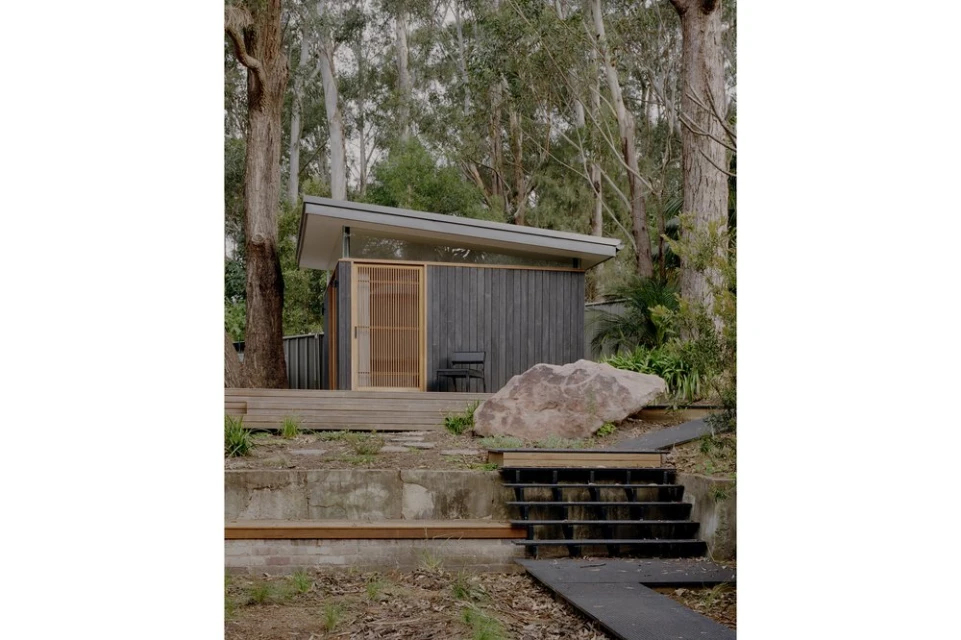 Architectural shot of a modern wooden cabin with sliding door, stone, and trees in a wooded area, evoking tranquil natural living.
