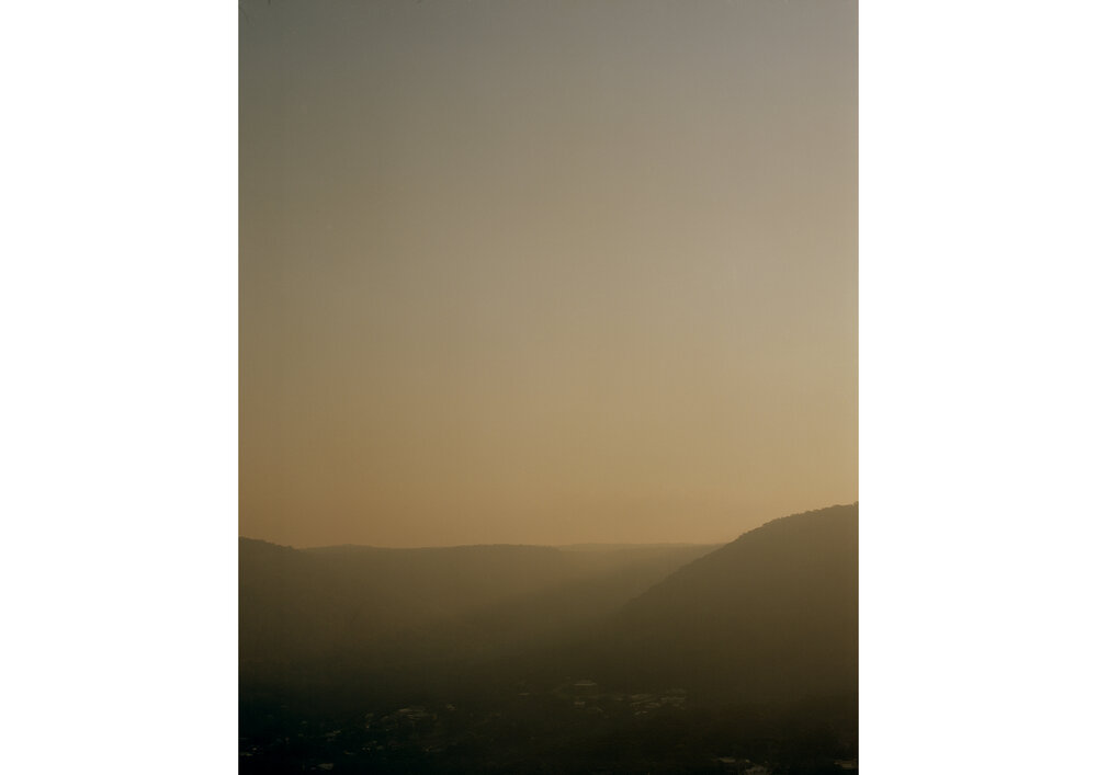 Golden hour view of mountain silhouettes with hazy skies. Warm lighting creates a tranquil, scenic landscape with a soft, muted palette.