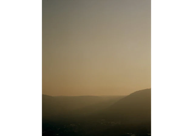 Golden hour view of mountain silhouettes with hazy skies. Warm lighting creates a tranquil, scenic landscape with a soft, muted palette.