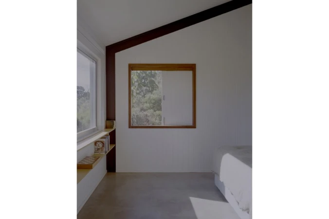 Interior bedroom view featuring a wooden framed window, floating shelf with books, and sloped ceiling by Daniel Mulheran