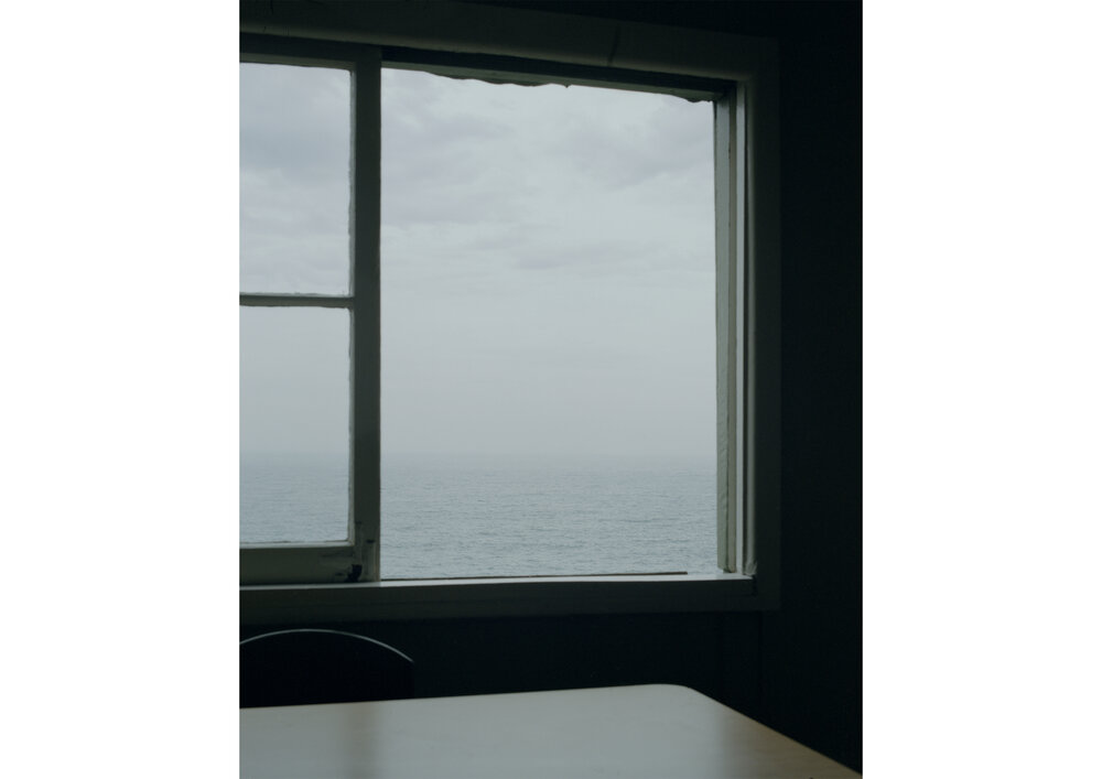 Indoor view of ocean through a window with a white frame, casting soft light onto a table and chair in a shadowed room.