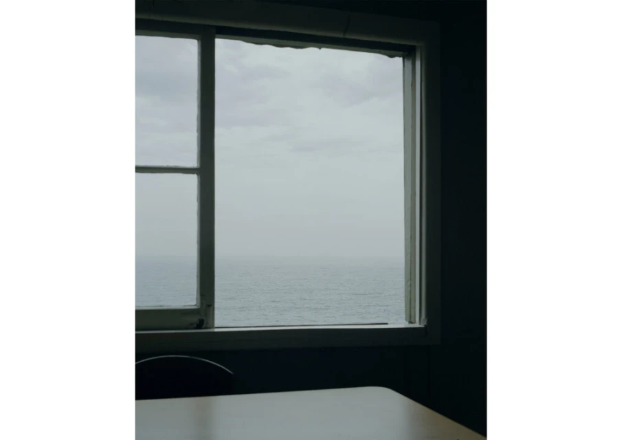 Indoor view of ocean through a window with a white frame, casting soft light onto a table and chair in a shadowed room.