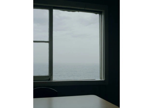 Indoor view of ocean through a window with a white frame, casting soft light onto a table and chair in a shadowed room.