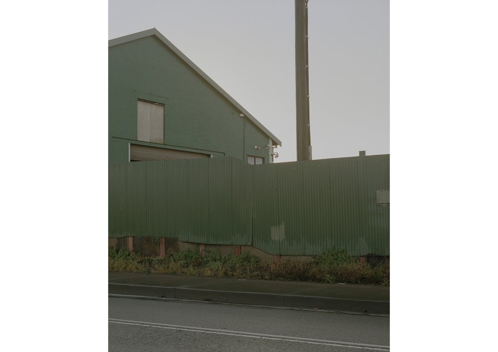 Exterior view of an industrial green building with a corrugated metal fence, featuring a concrete base and an overcast sky.