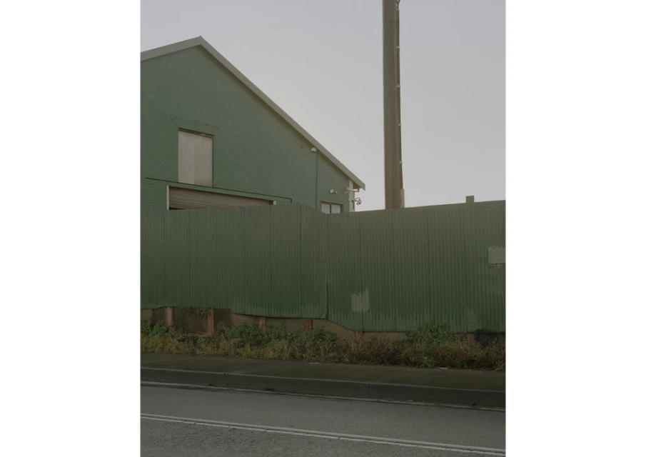 Exterior view of an industrial green building with a corrugated metal fence, featuring a concrete base and an overcast sky.