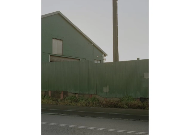 Exterior view of an industrial green building with a corrugated metal fence, featuring a concrete base and an overcast sky.