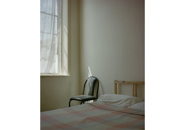 Sunlit corner of a minimalist bedroom with a bed, metal chair, and window with sheer white curtains.
