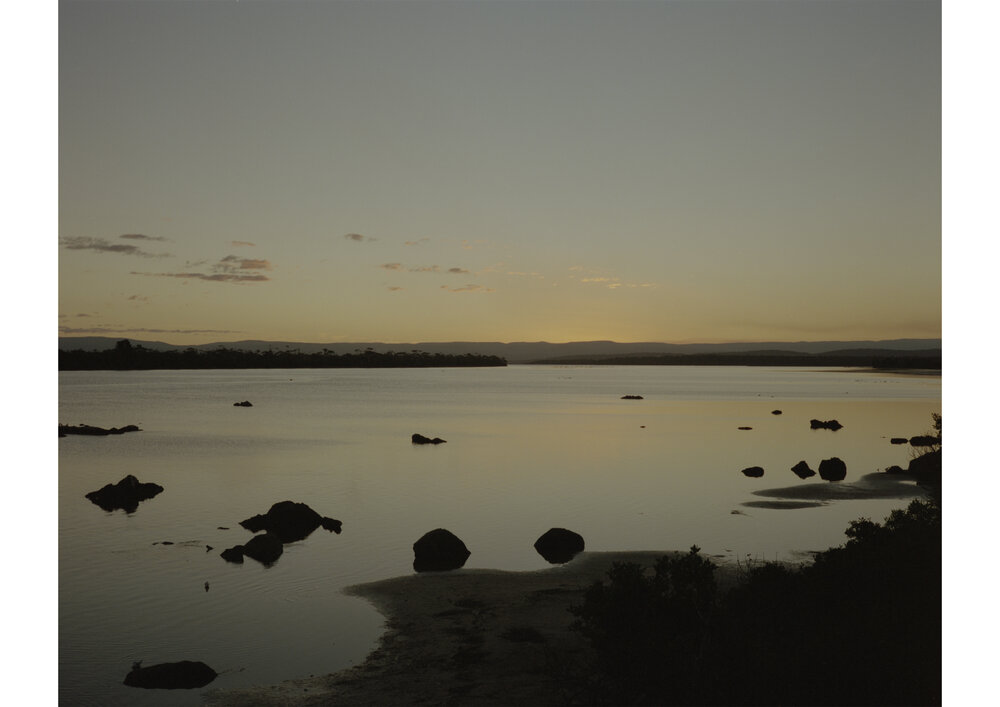Scenic dusk view of a tranquil lake with scattered rocks and distant trees under a muted, warm-toned sky, creating a peaceful landscape.