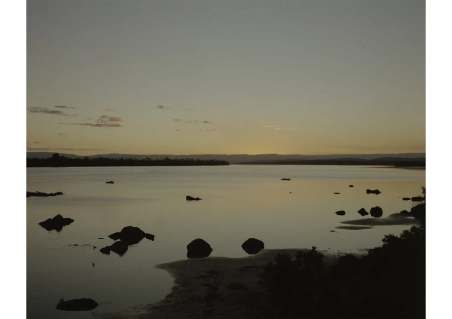 Scenic dusk view of a tranquil lake with scattered rocks and distant trees under a muted, warm-toned sky, creating a peaceful landscape.
