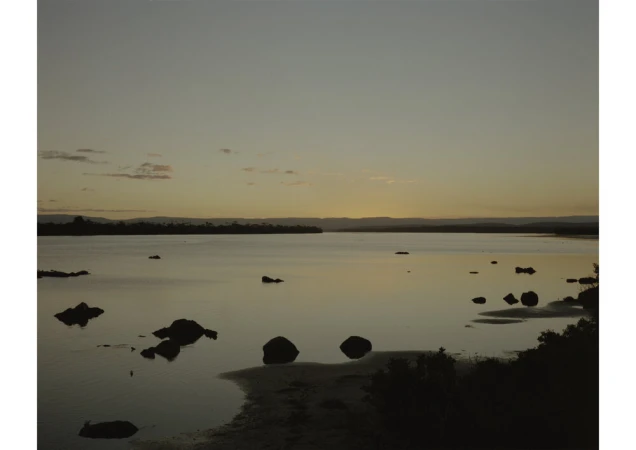 Scenic dusk view of a tranquil lake with scattered rocks and distant trees under a muted, warm-toned sky, creating a peaceful landscape.