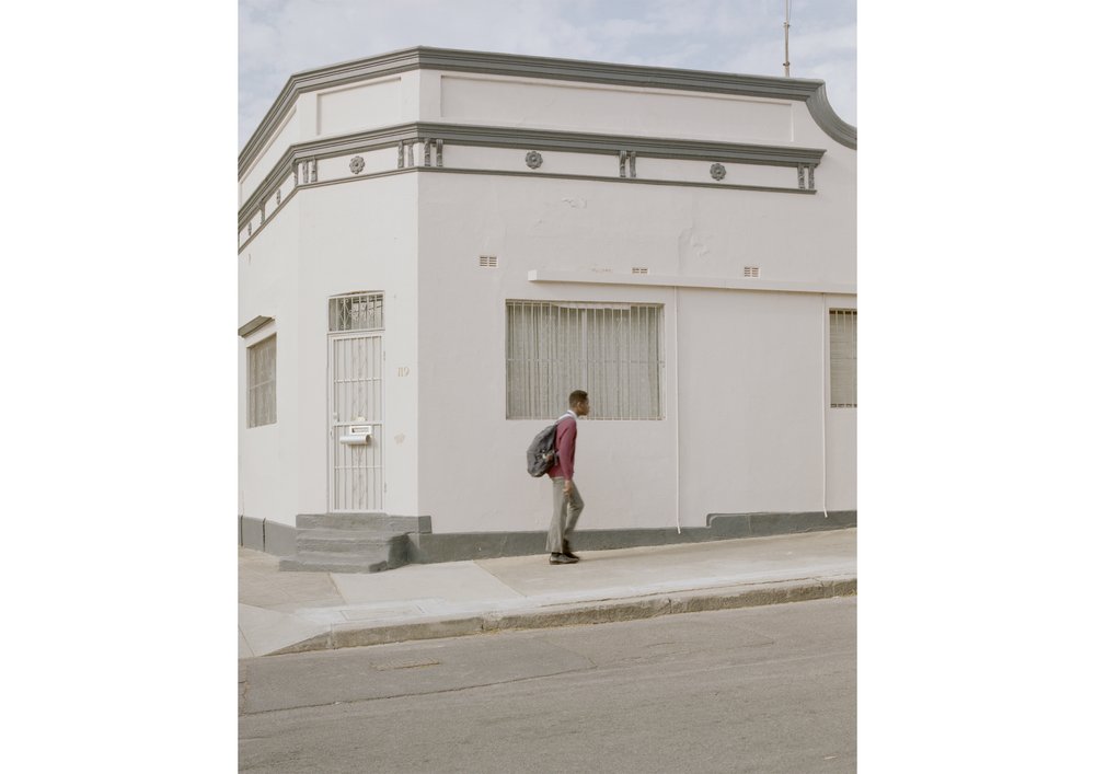 Young adult walks on a sidewalk next to a white corner building on a sunny day, wearing a backpack and casual clothing.