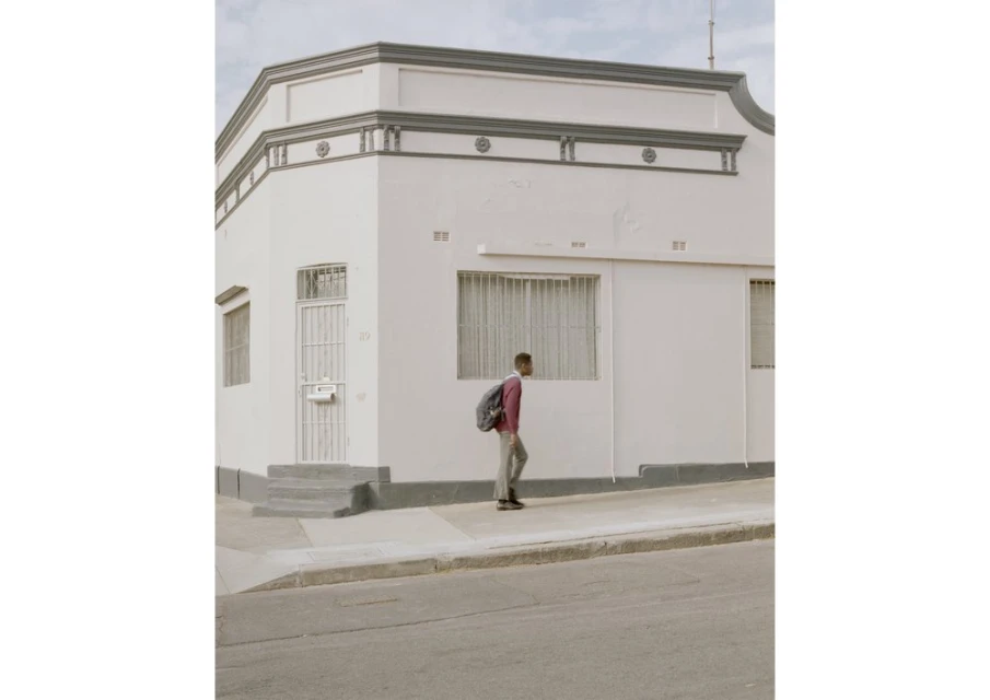 Young adult walks on a sidewalk next to a white corner building on a sunny day, wearing a backpack and casual clothing.