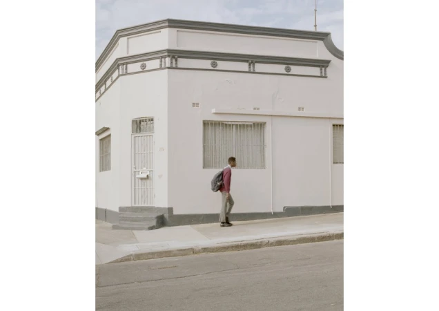 Young adult walks on a sidewalk next to a white corner building on a sunny day, wearing a backpack and casual clothing.
