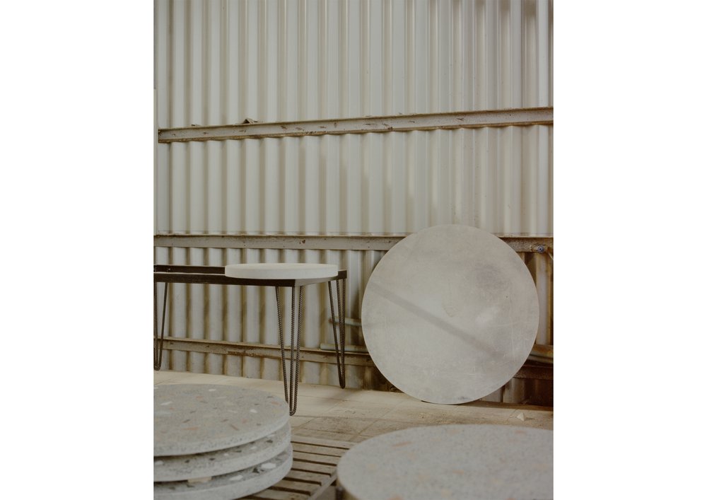 Minimalist interior with round concrete coffee table and side table in a neutral color palette, set against a corrugated metal wall.