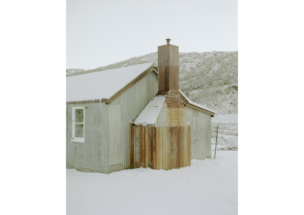 Exterior view of a corrugated metal building covered in snow in a winter landscape with a stone chimney and hillside backdrop.