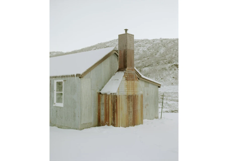 Exterior view of a corrugated metal building covered in snow in a winter landscape with a stone chimney and hillside backdrop.