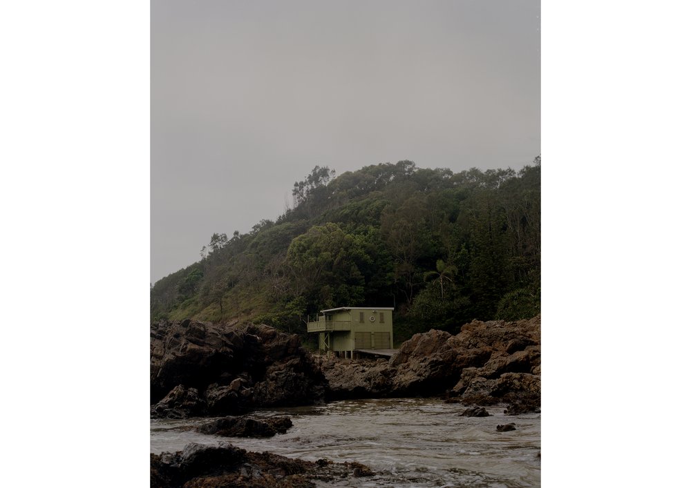 Coastal view of a small house nestled among rocks and lush vegetation under an overcast sky, capturing a serene, secluded scene.