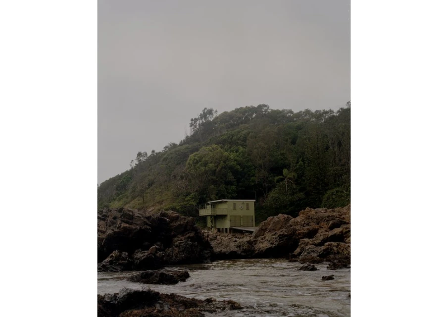Coastal view of a small house nestled among rocks and lush vegetation under an overcast sky, capturing a serene, secluded scene.