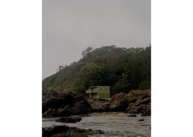 Coastal view of a small house nestled among rocks and lush vegetation under an overcast sky, capturing a serene, secluded scene.