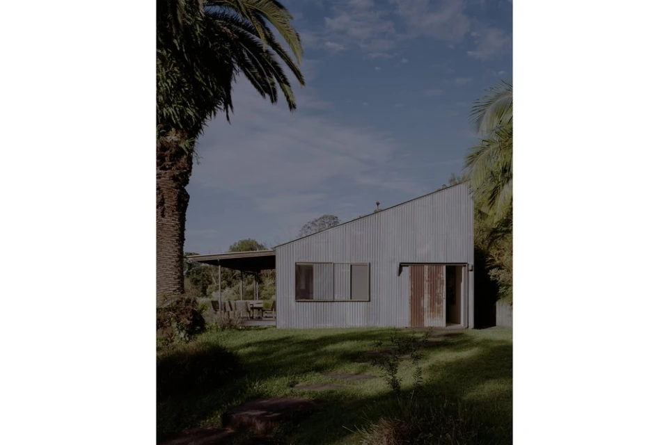 Exterior view of a modern corrugated metal house with large windows and a covered porch, surrounded by lush greenery and palm trees.