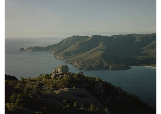 Aerial shot of rocky cliffs meeting the ocean, with green, hilly mountains in the background on a sunny day. Nature scene, landscape.