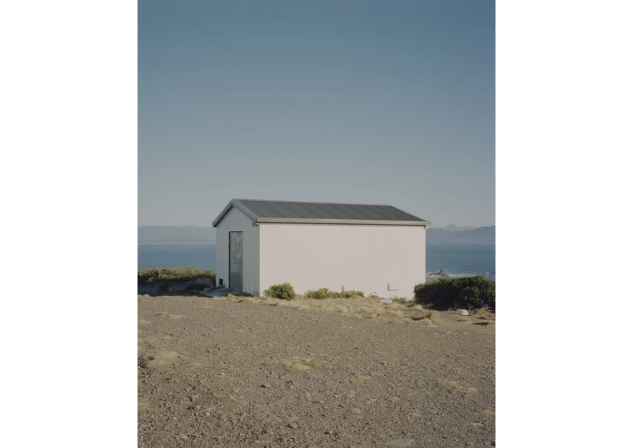 Exterior view of a small, white, rectangular building with a gray roof, standing on a rocky hill overlooking the ocean and a clear blue sky.