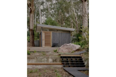 Modern dark timber cabin with a slanted roof nestled among tall eucalyptus trees and a boulder in a natural bushland setting