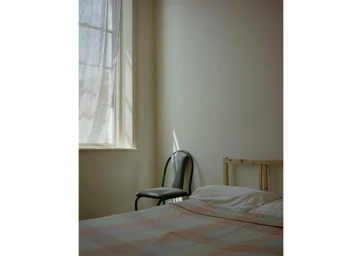 Sunlit corner of a minimalist bedroom with a bed, metal chair, and window with sheer white curtains.