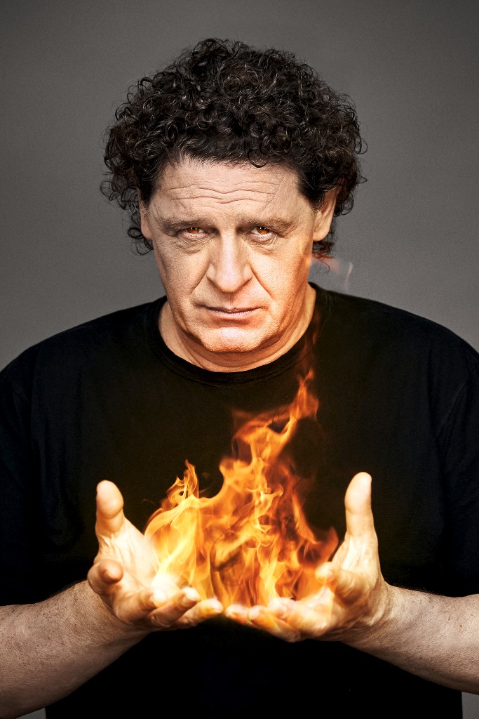 Portrait of an adult male with dark curly hair, wearing a black T-shirt and holding fire in his hands against a grey backdrop.