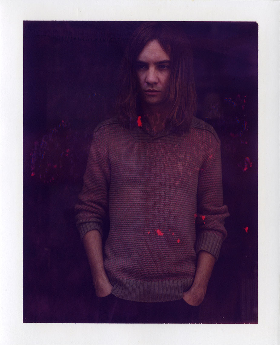 Portrait of a young adult male with shoulder-length brown hair, wearing a brown sweater, standing indoors against a dark background.