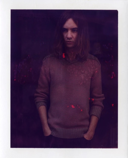 Portrait of a young adult male with shoulder-length brown hair, wearing a brown sweater, standing indoors against a dark background.