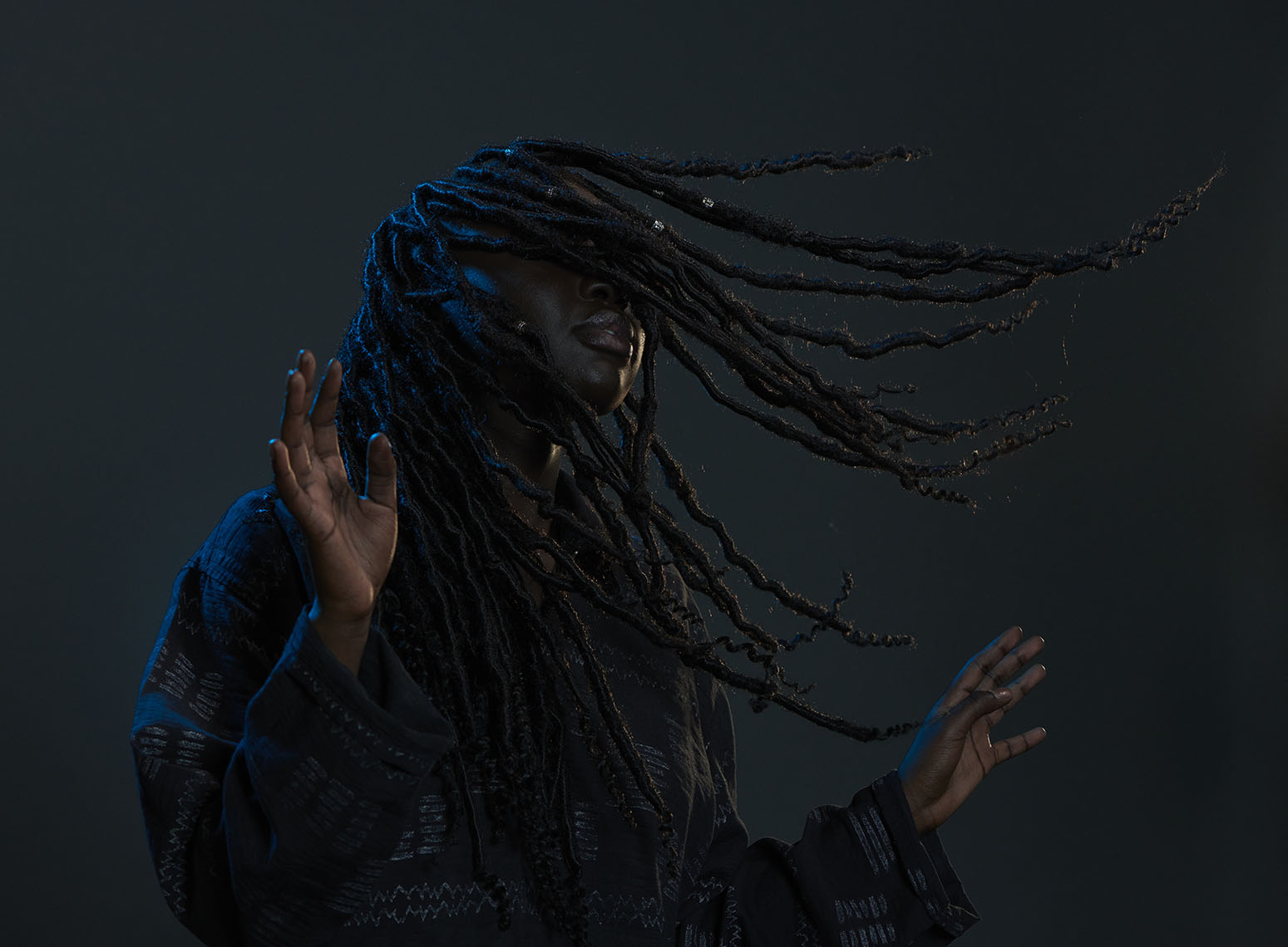 Black woman with dreadlocks flowing across her face wearing a dark patterned shirt against a dark background, photographed by Ian & Erick.